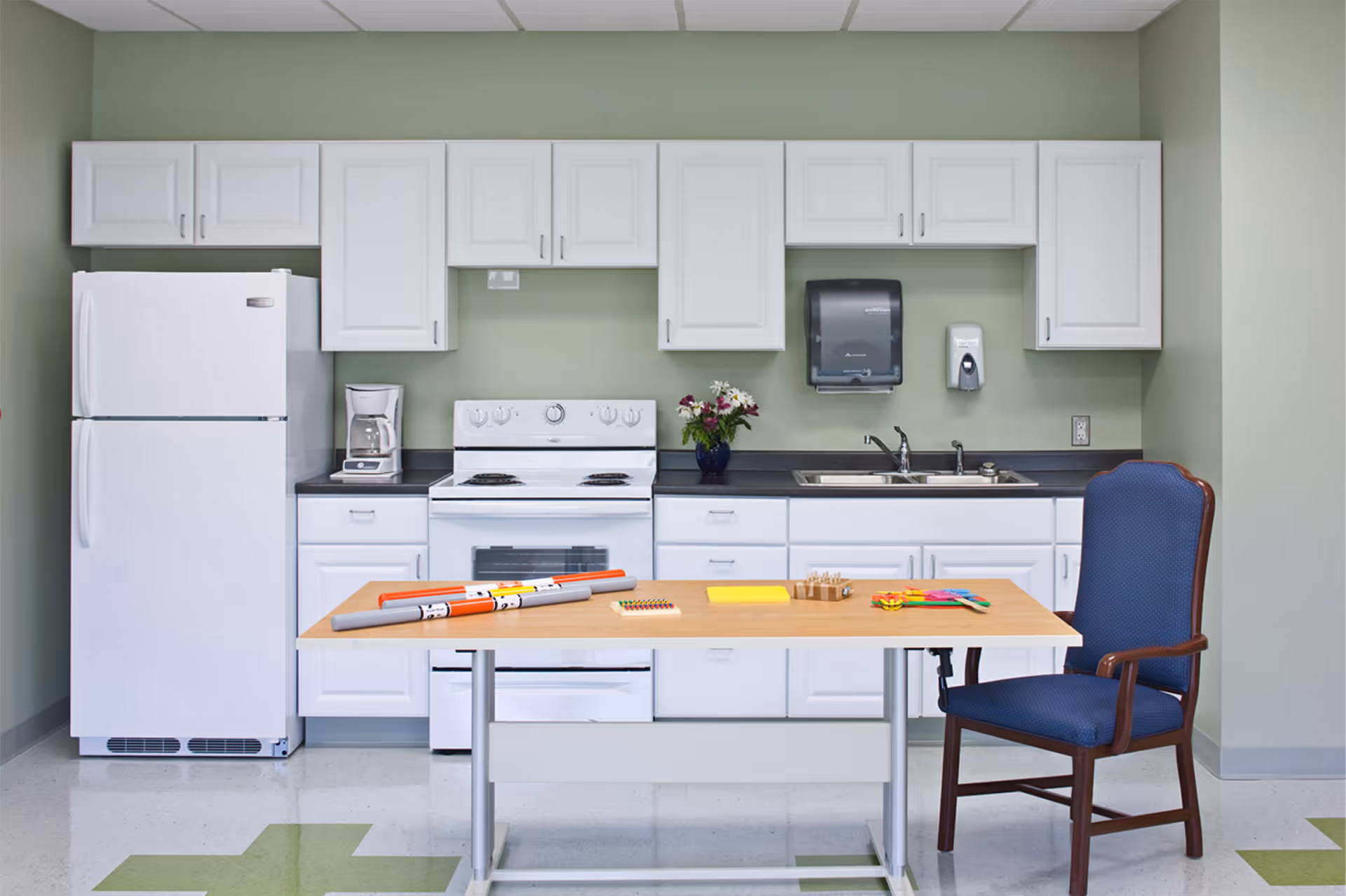 A clean and organized kitchen area with white cabinets, a white refrigerator, stove, and coffee maker. A wooden table with various colorful items and a blue cushioned chair are in the foreground. The walls are painted light green and there is a paper towel dispenser and soap dispenser mounted above the sink.