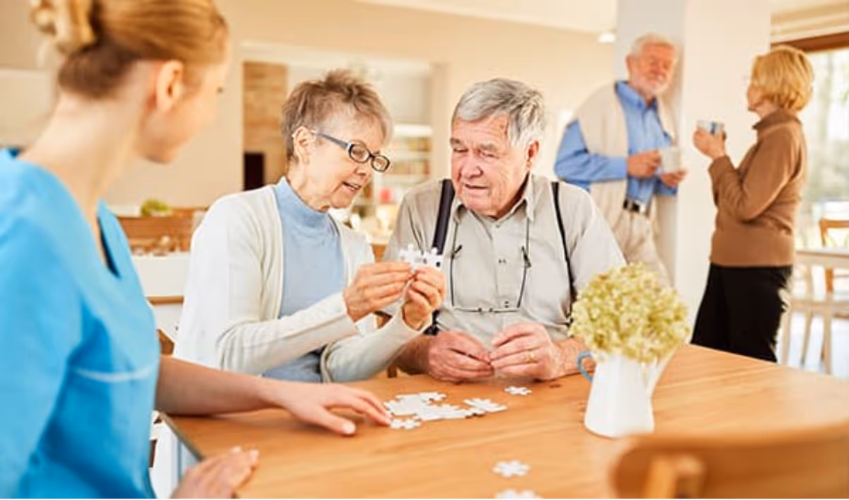 Two elderly individuals sitting at a table working on a jigsaw puzzle with a caregiver in blue scrubs watching them. In the background, another elderly couple is standing and having a conversation while holding cups. The setting appears to be a bright and welcoming common area in a senior living facility.