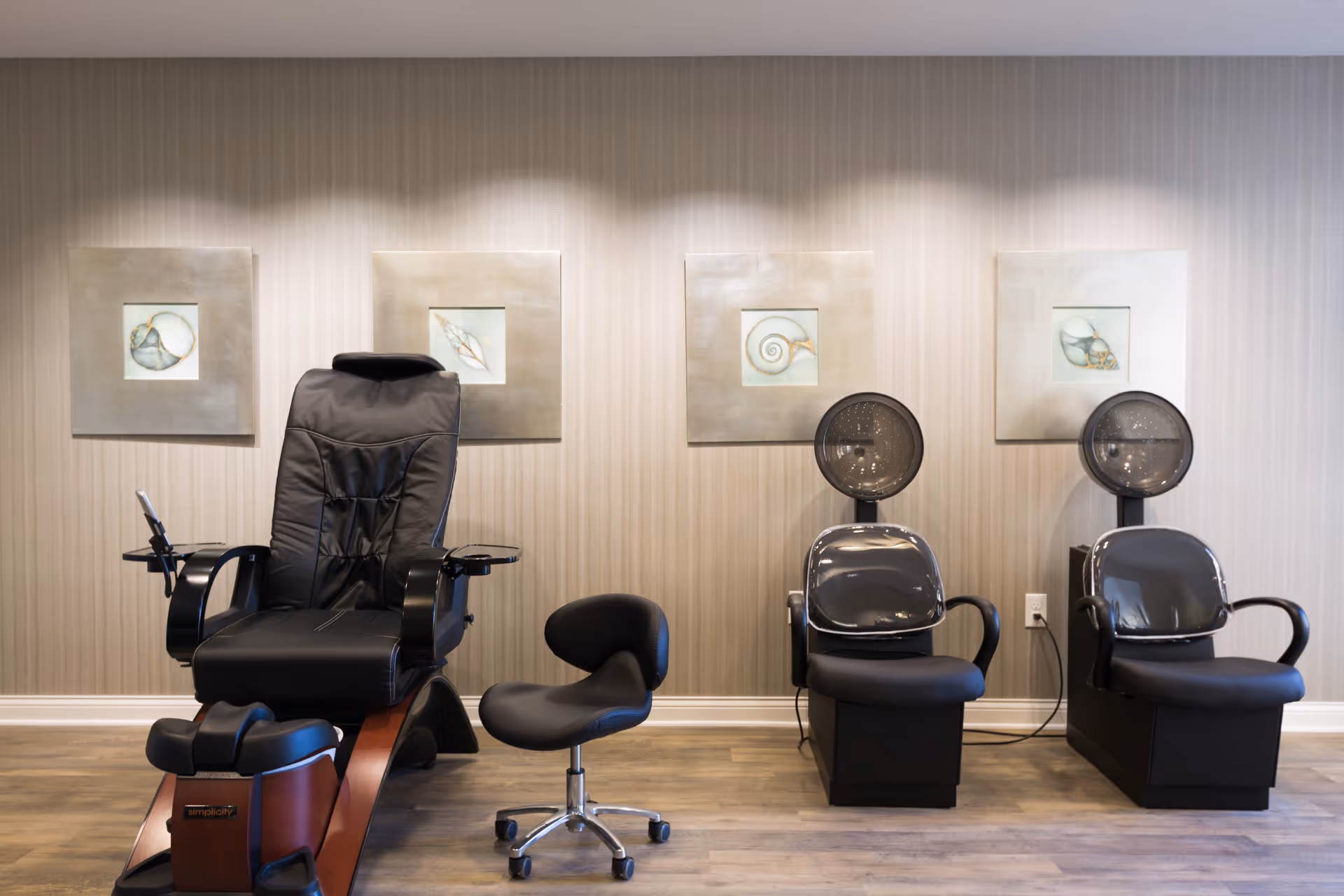 A salon area with a black pedicure chair, a small black rolling stool, and two black hair dryer chairs against a beige striped wall with four framed pictures of seashells.