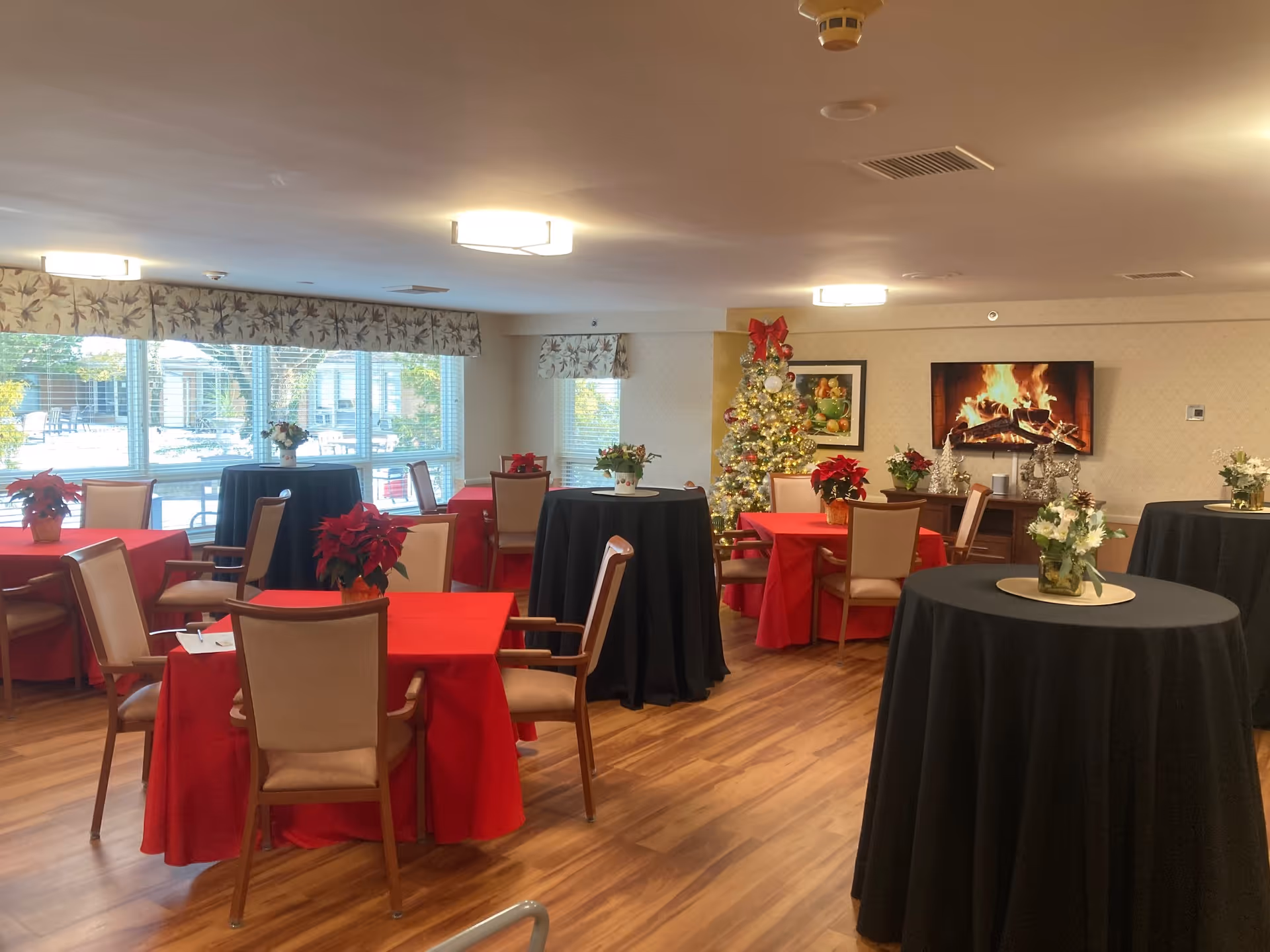 Communal dining room decorated for Christmas with tables and chairs, poinsettias, a decorated tree, and a TV showing a fireplace.