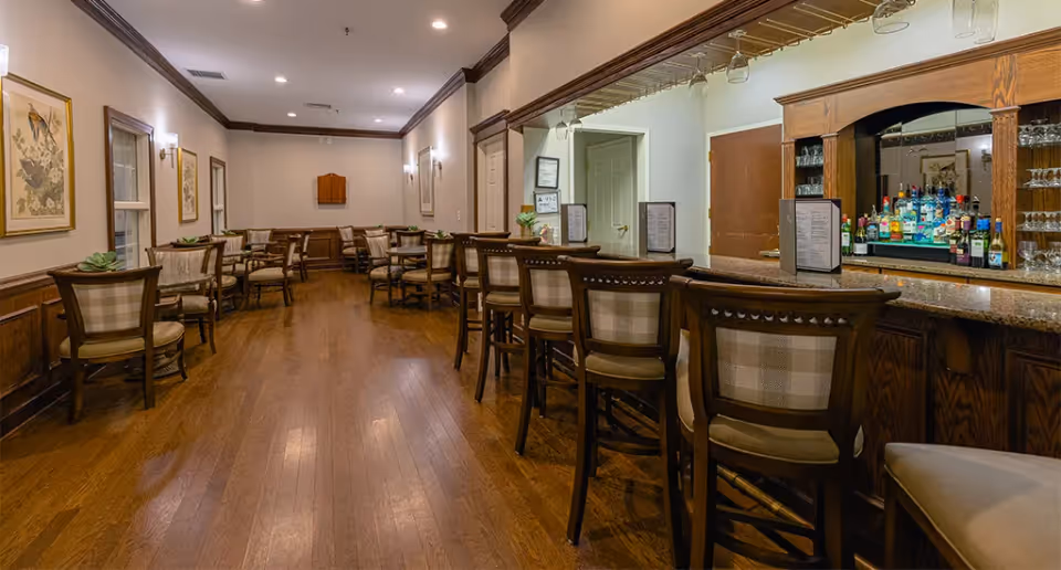 Dining room and bar lounge with wooden floors, a row of barstools, and several small tables and chairs.