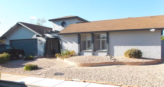 Exterior view of a single-story residential building with a brown shingle roof, light gray walls, and a blue garage door. The front yard is landscaped with gravel and a few small bushes. A black vehicle is parked in the driveway.