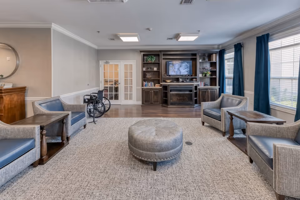 A spacious living room with four gray armchairs arranged around a round gray ottoman on a large carpet. There are two wooden side tables next to the chairs. A dark wooden entertainment center with a TV and shelves is against the far wall. Large windows with blue curtains let in natural light. A wheelchair is positioned near a set of white French doors.