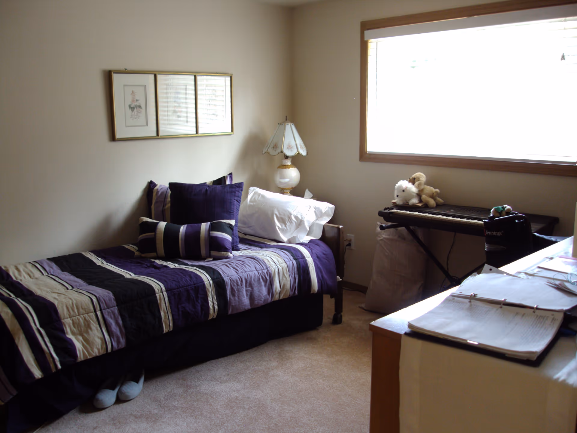 A bedroom with a single bed covered in a purple, black, and beige striped comforter and multiple pillows. There is a bedside table with a decorative lamp next to the bed. A keyboard with stuffed animals on top is placed near a large window with blinds. A desk with open binders and papers is visible in the foreground.