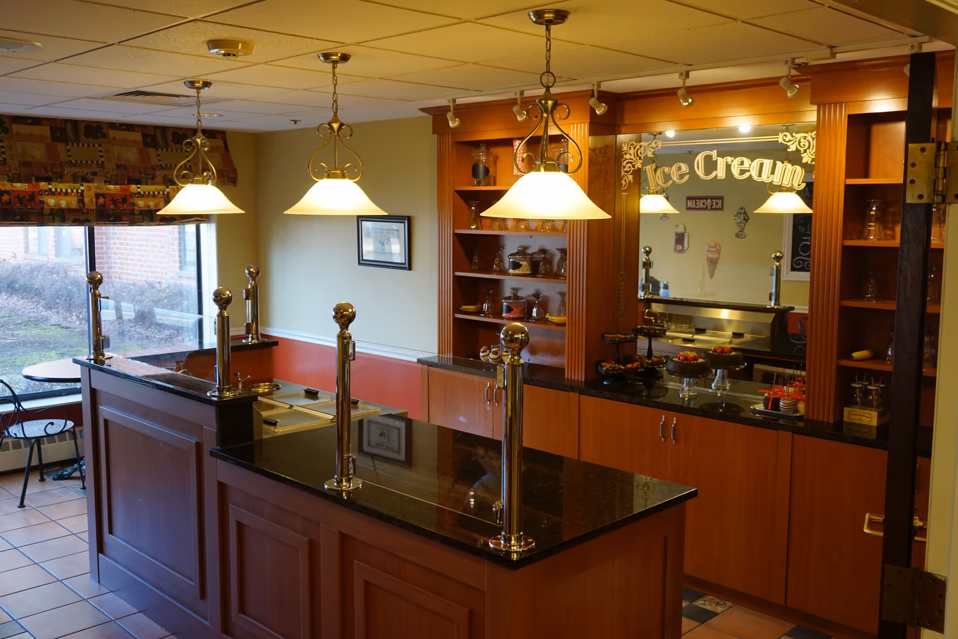 An ice cream serving area inside a facility with a wooden counter and black granite countertop. There are three hanging pendant lights above the counter. Behind the counter, there are shelves with glass jars and a mirror with the words 'Ice Cream' written on it. On the counter, there are various desserts displayed. A window with a patterned valance and a small table with chairs are visible to the left.