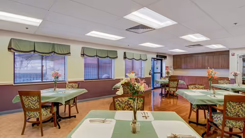 A dining room in a senior living facility with several tables covered in green and white tablecloths, each set with utensils and a vase of flowers. The room has large windows with green valances, wooden chairs with patterned cushions, and a wooden cabinet area in the back near the entrance door.