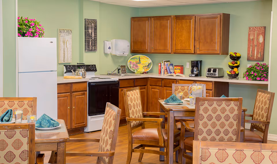A cozy dining area with wooden tables and cushioned chairs featuring patterned upholstery. In the background, there is a kitchen with wooden cabinets, a white refrigerator, a black stove, a coffee maker, a toaster, and decorative items including plants and wall art with kitchen and dining themes. The walls are painted light green.