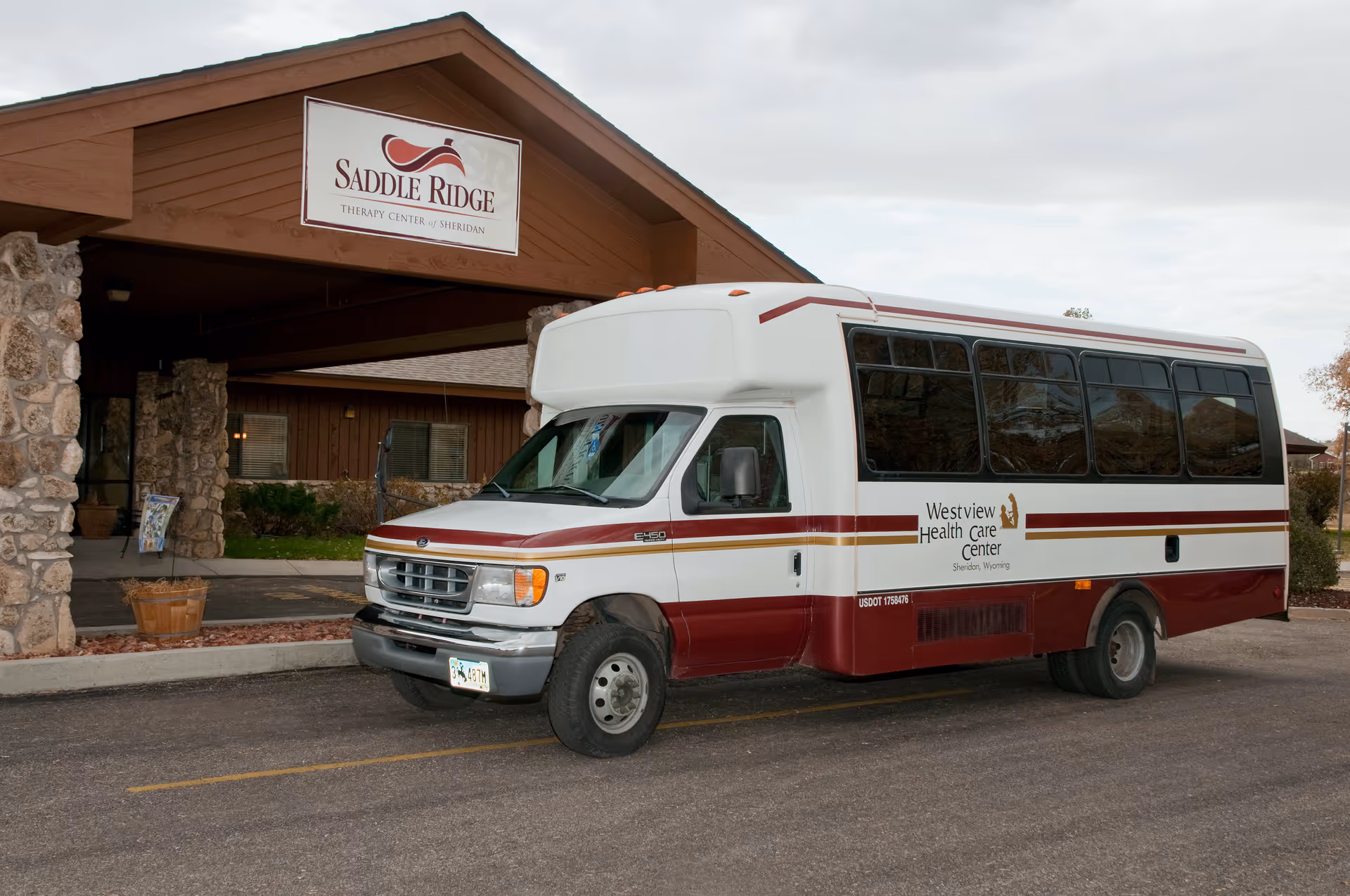 A white and maroon shuttle bus parked in front of a building entrance with a sign that reads 'Saddle Ridge Therapy Center of Sheridan'. The bus has 'Westview Health Care Center Sheridan, Wyoming' written on its side.