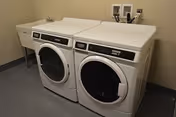 A laundry room with a white front-loading washing machine and dryer side by side, a utility sink to the left, and a beige wall with electrical outlets above the machines.