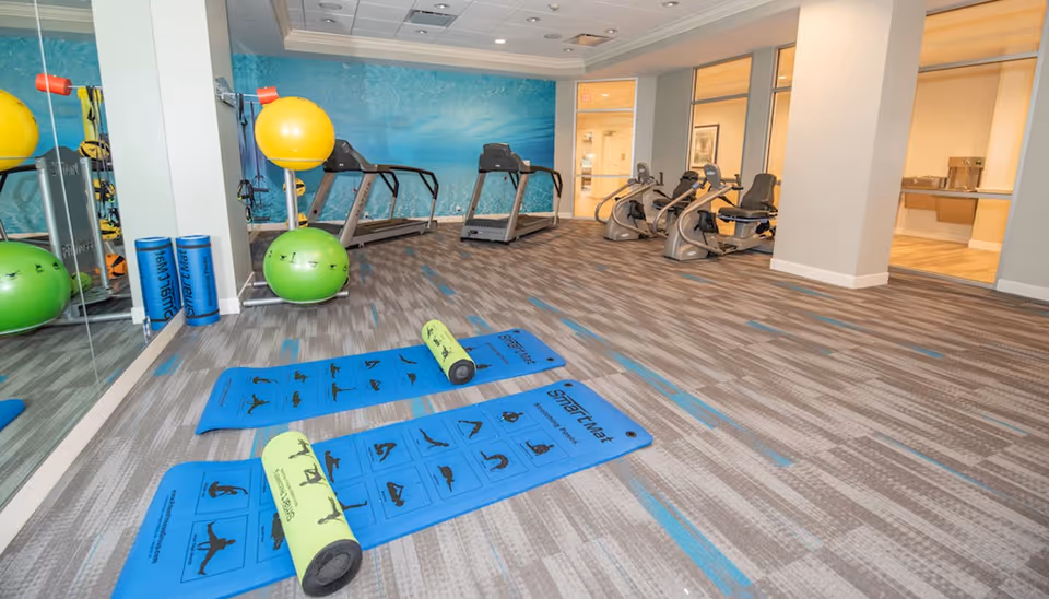 Fitness room with blue exercise mats and foam rollers on the floor, stability balls, treadmills and recumbent bikes against a mirrored wall.