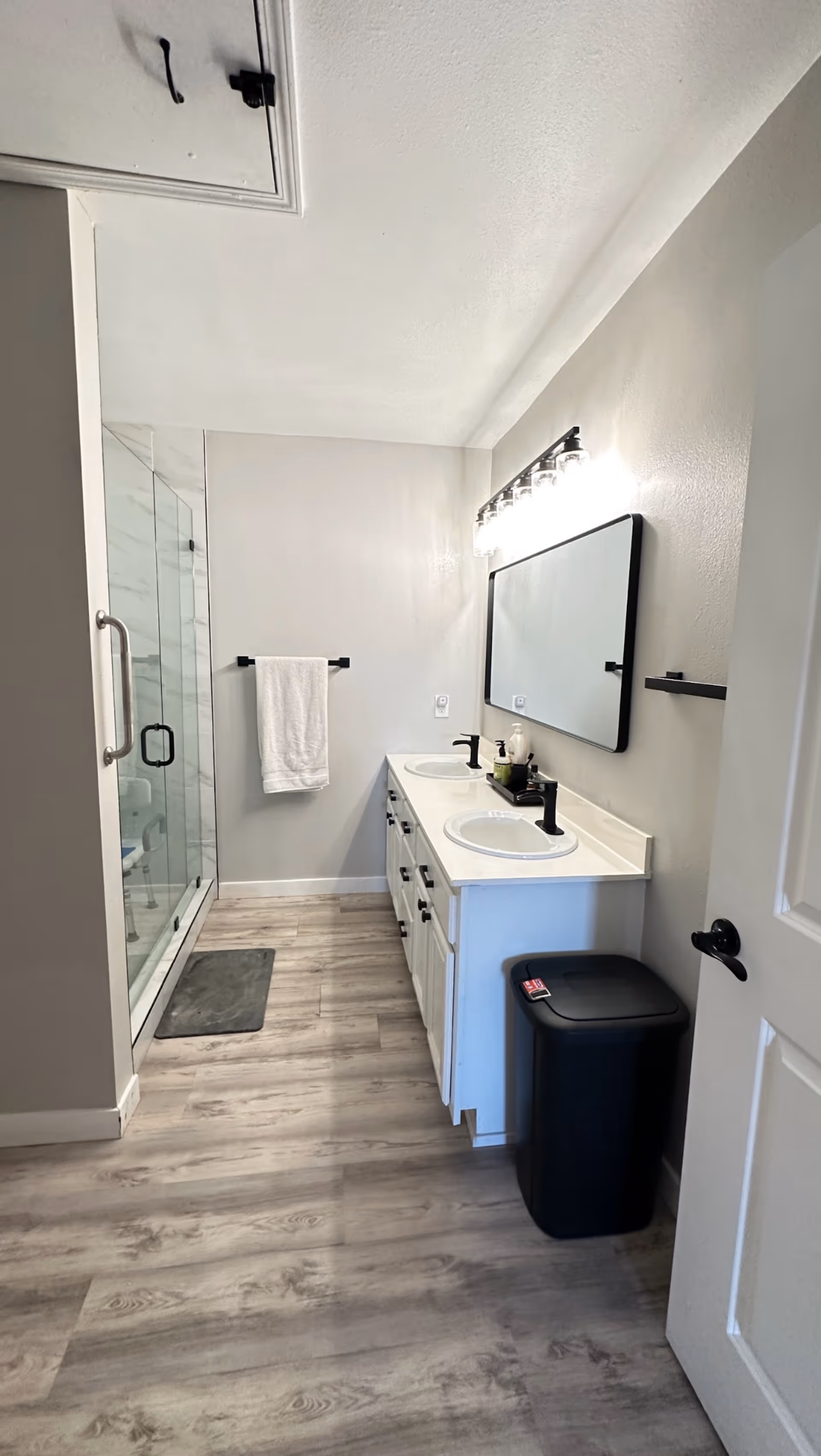 A modern bathroom with a double sink vanity featuring black faucets and a large rectangular mirror above. To the left, there is a glass-enclosed shower with a towel hanging on a black towel bar. The floor has light wood-style flooring, and a black trash bin is placed near the white door on the right.