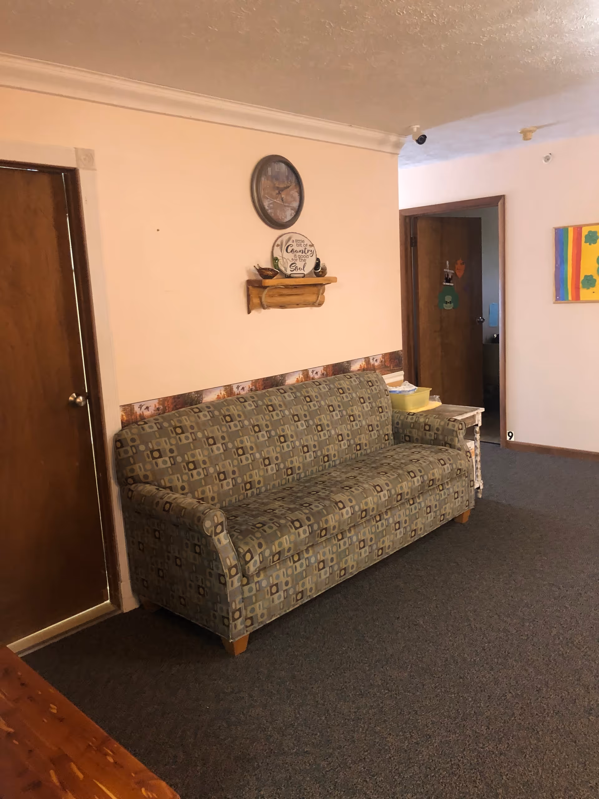 A living room area with a patterned sofa against a light pink wall. Above the sofa is a round clock and a small wooden shelf holding decorative items. There is a wooden door to the left and an open doorway to the right, with a colorful painting on the far right wall. The floor is carpeted in a dark color.