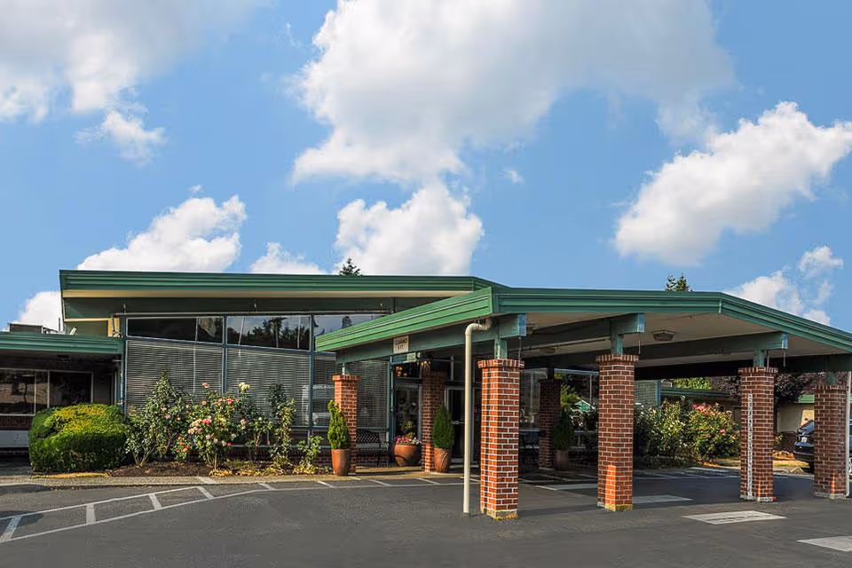 Exterior view of Life Care Center of Puyallup showing the building entrance with a covered drop-off area supported by brick columns, surrounded by landscaped bushes and flowers under a partly cloudy sky.