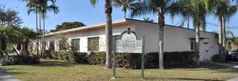 Single-story white senior living facility with palm trees and a visible front sign reading Homestead Manor.