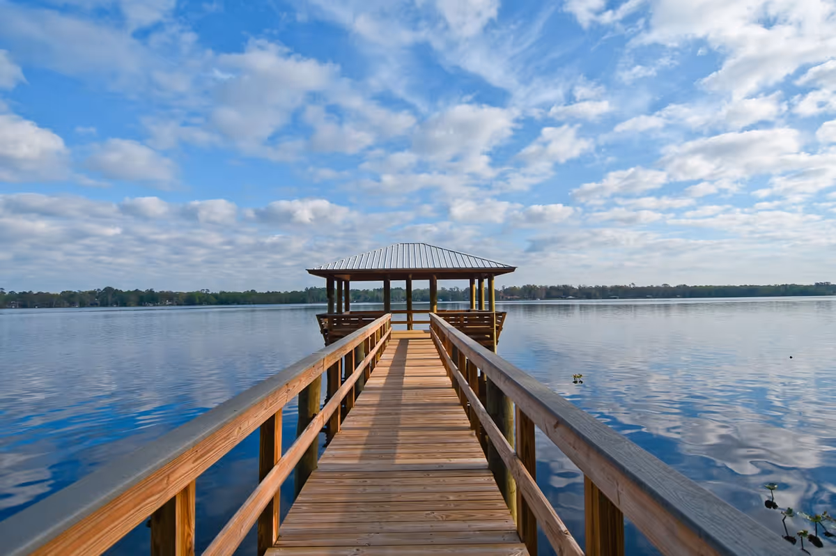 A wooden pier leading out over a calm lake with a covered gazebo at the end, under a partly cloudy blue sky.