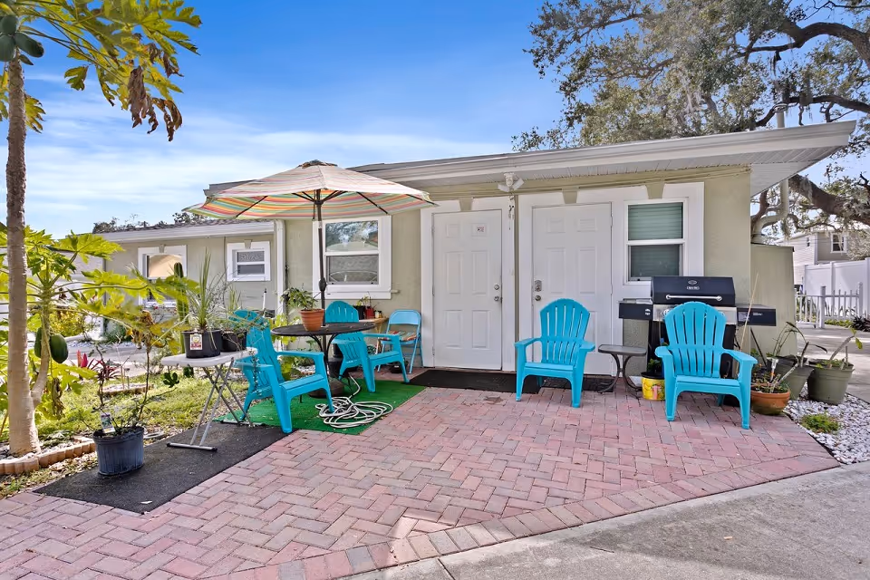Outdoor patio area with a small table and four turquoise chairs under a striped umbrella. There are potted plants around the patio and a barbecue grill next to the building with two white doors and a window. The area is paved with red bricks and surrounded by greenery and trees.