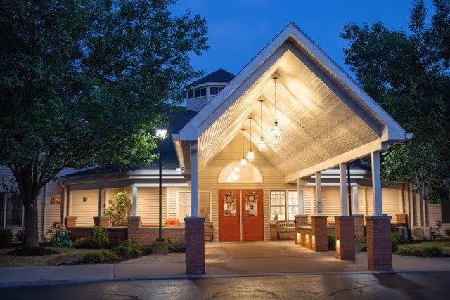 Entrance of Ashton Place Senior Living at dusk with a covered driveway supported by brick pillars, warm hanging lights, red double doors decorated with wreaths, and surrounding trees and landscaping.