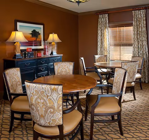 A cozy dining area with two round wooden tables surrounded by upholstered chairs with patterned backs and tan seats. The room features warm brown walls, a patterned carpet, a window with curtains, and a black sideboard with two table lamps and decorative items including large dice and a framed landscape picture.