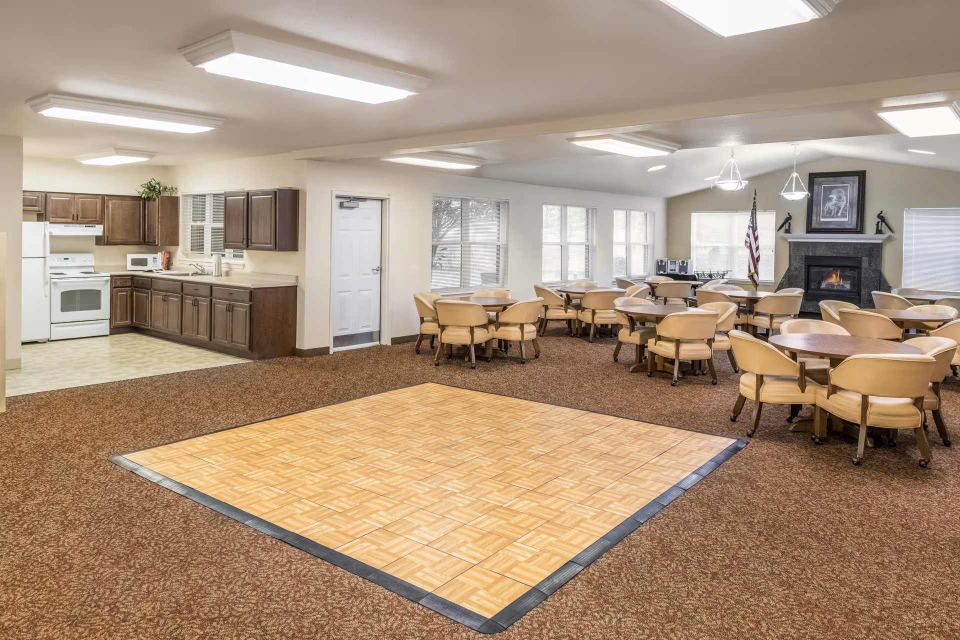 A spacious room featuring a kitchen area with dark wood cabinets, a white stove, microwave, and refrigerator on the left side. The room also includes multiple round tables with beige chairs arranged on a carpeted floor, and a small square wooden dance floor in the center. Large windows allow natural light to enter, and a fireplace with an American flag and framed artwork is visible in the background.