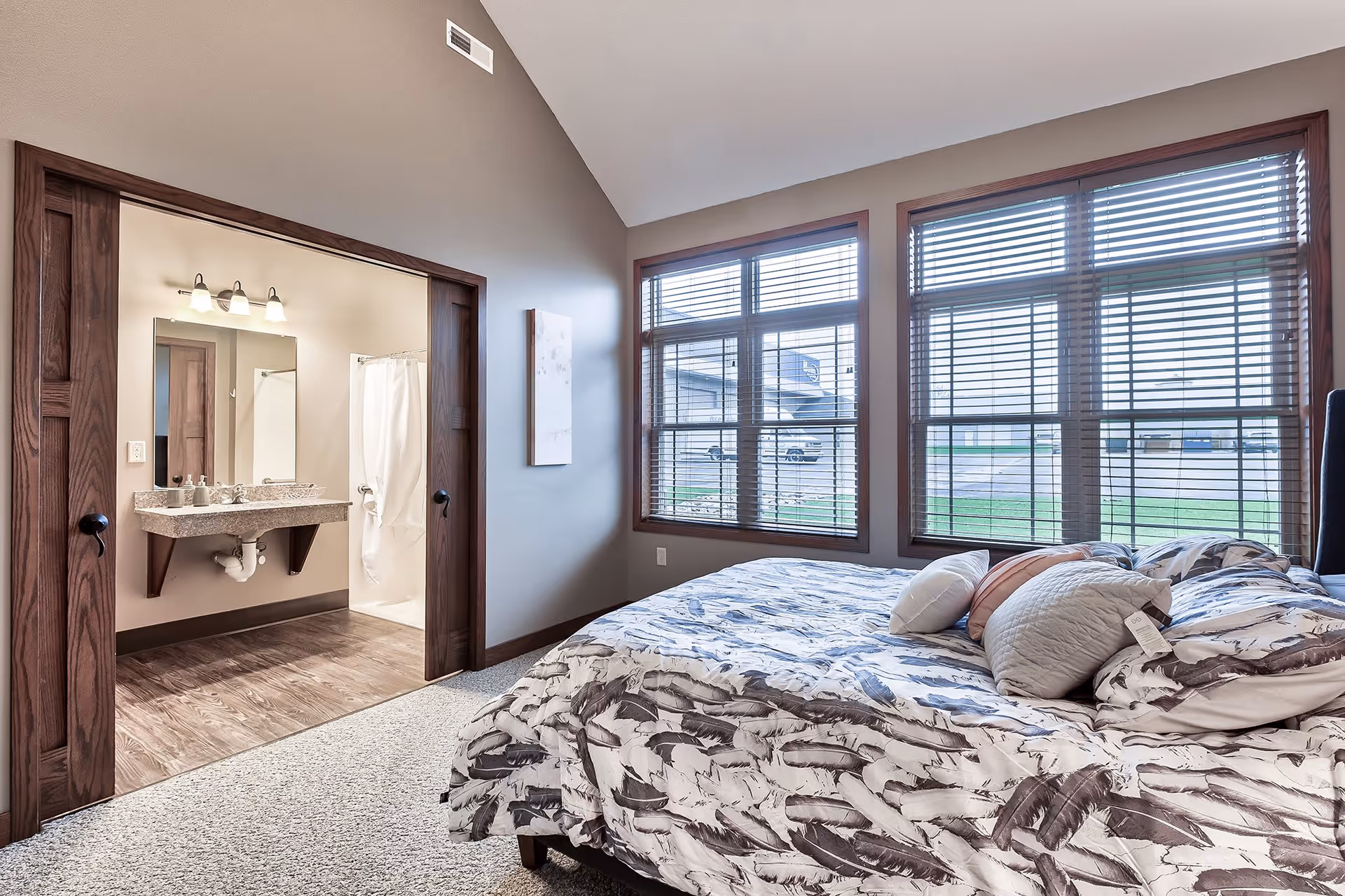 A bedroom with a bed covered in a feather-patterned comforter and several pillows. The room has large windows with wooden blinds letting in natural light. To the left, there is an open doorway leading to a bathroom with a granite countertop sink and a shower with a white curtain.