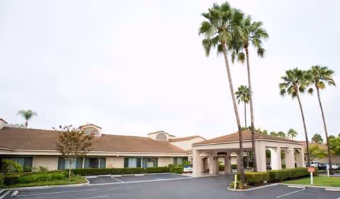 Single-story senior living facility exterior with driveway, palm trees, and covered entrance.