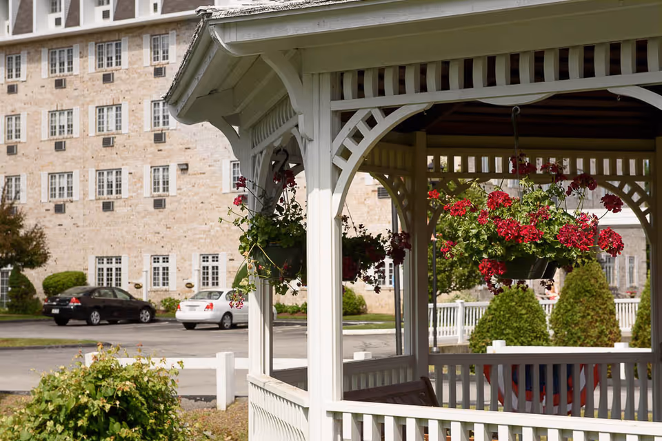 White wooden gazebo with hanging red flower baskets in front of a multi-story brick senior living building.