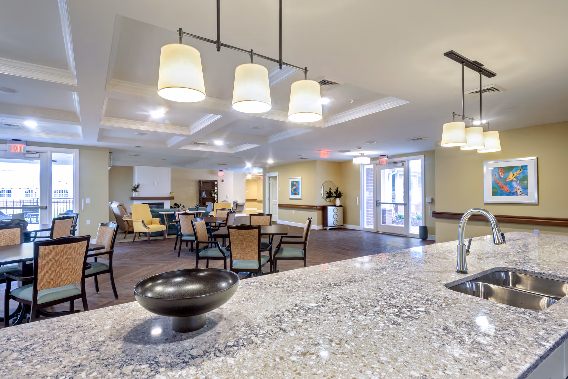 A spacious and well-lit common area in a senior living facility featuring multiple tables and chairs arranged for dining or socializing. The foreground shows a granite countertop with a modern sink and a decorative bowl. The room has soft yellow walls, ceiling lights with lampshades, framed artwork, and large glass doors leading to an outdoor area.