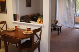 Small dining area with a wooden table set for two next to a pass-through into a carpeted living room with a sliding glass door to a balcony.
