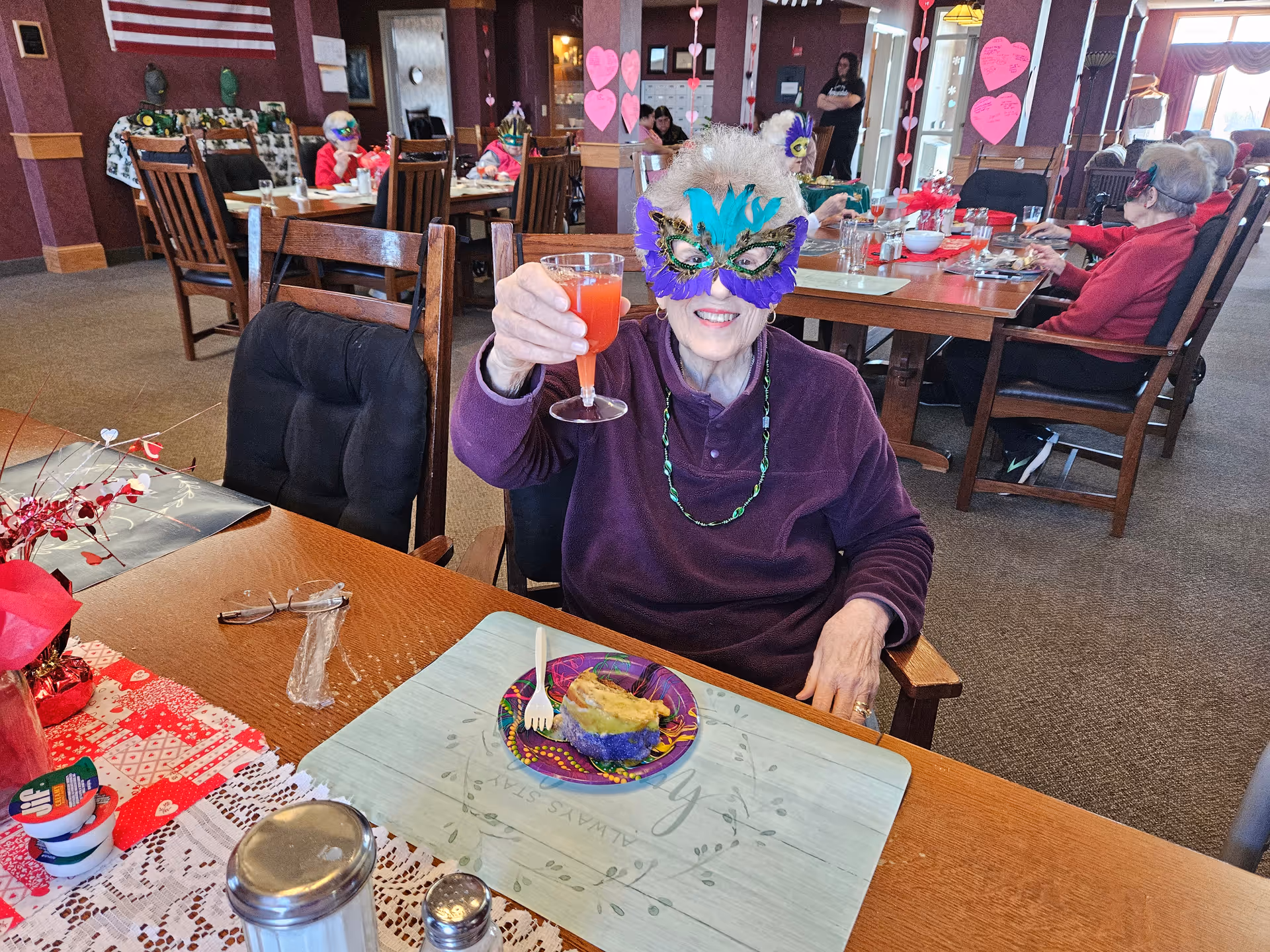 An elderly woman wearing a colorful feathered mask and a purple sweater is sitting at a dining table in a communal dining room. She is holding up a glass of red drink and smiling. The table has a plate with a piece of cake, a fork, and some condiments. Other elderly people, also wearing masks, are seated at tables in the background. The room is decorated with heart-shaped paper cutouts hanging from the ceiling and an American flag on the wall.