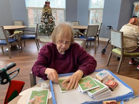An elderly woman wearing a maroon sweater is sitting at a table in a common area, engaging in a craft activity with holiday-themed materials. Behind her, there is a decorated Christmas tree near large windows, and other chairs and tables are visible in the room.