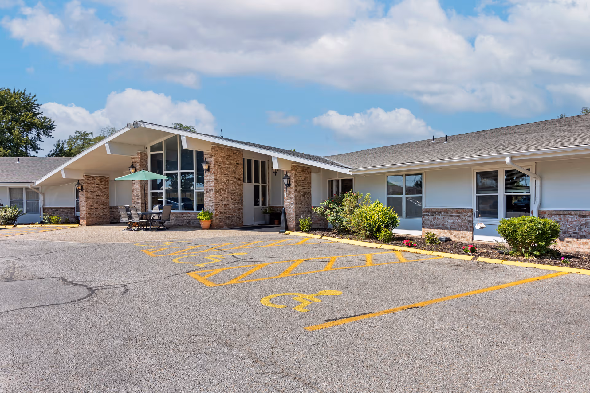 Single-story brick and siding senior living facility front entrance with a covered porte-cochere, outdoor seating with an umbrella, and marked parking spaces.