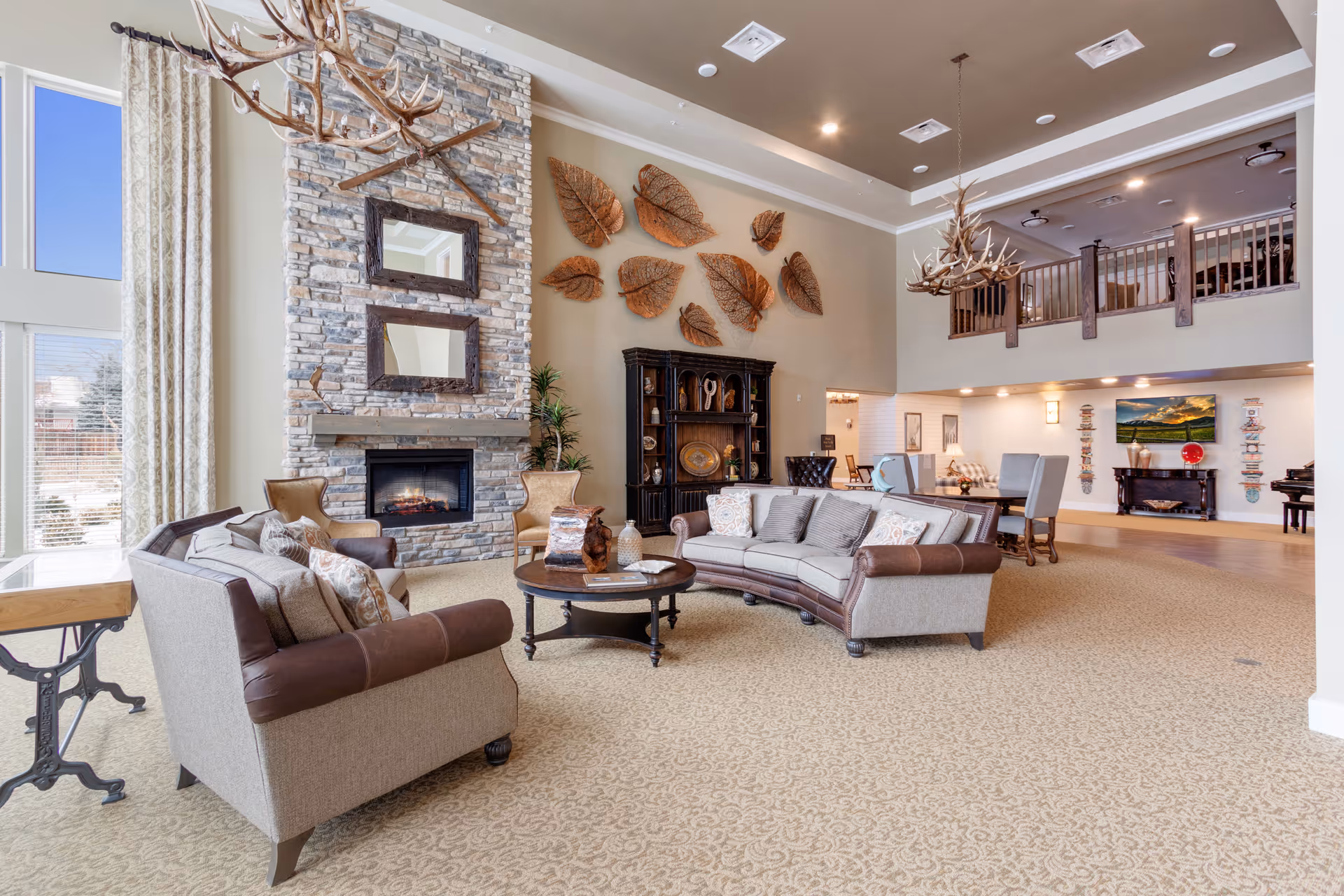 A spacious and well-lit living room area with beige carpet and high ceilings. The room features two sofas with brown leather accents, a round wooden coffee table, and two armchairs near a stone fireplace with two rectangular mirrors above it. Large windows with patterned curtains allow natural light to fill the space. Decorative antler chandeliers hang from the ceiling, and a wall is adorned with large leaf sculptures. In the background, there is a dining area with a table and chairs, a black cabinet, and a balcony railing on the upper level.