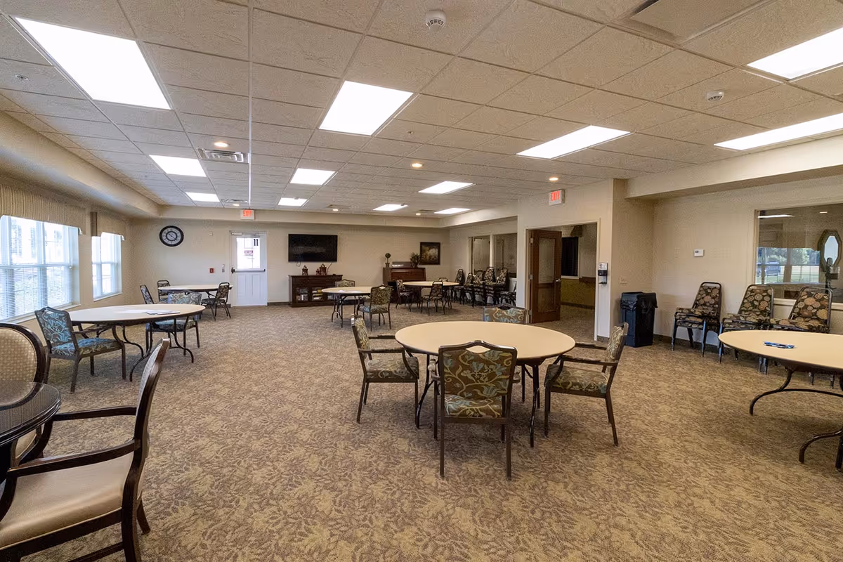 A spacious, well-lit common room with multiple round tables and chairs arranged around the room. The room has beige patterned carpet, white walls, and a drop ceiling with fluorescent lights. There is a wall clock, a flat-screen TV mounted on the far wall, and a door with an exit sign above it. Additional chairs are stacked along the walls, and windows on the left side let in natural light.