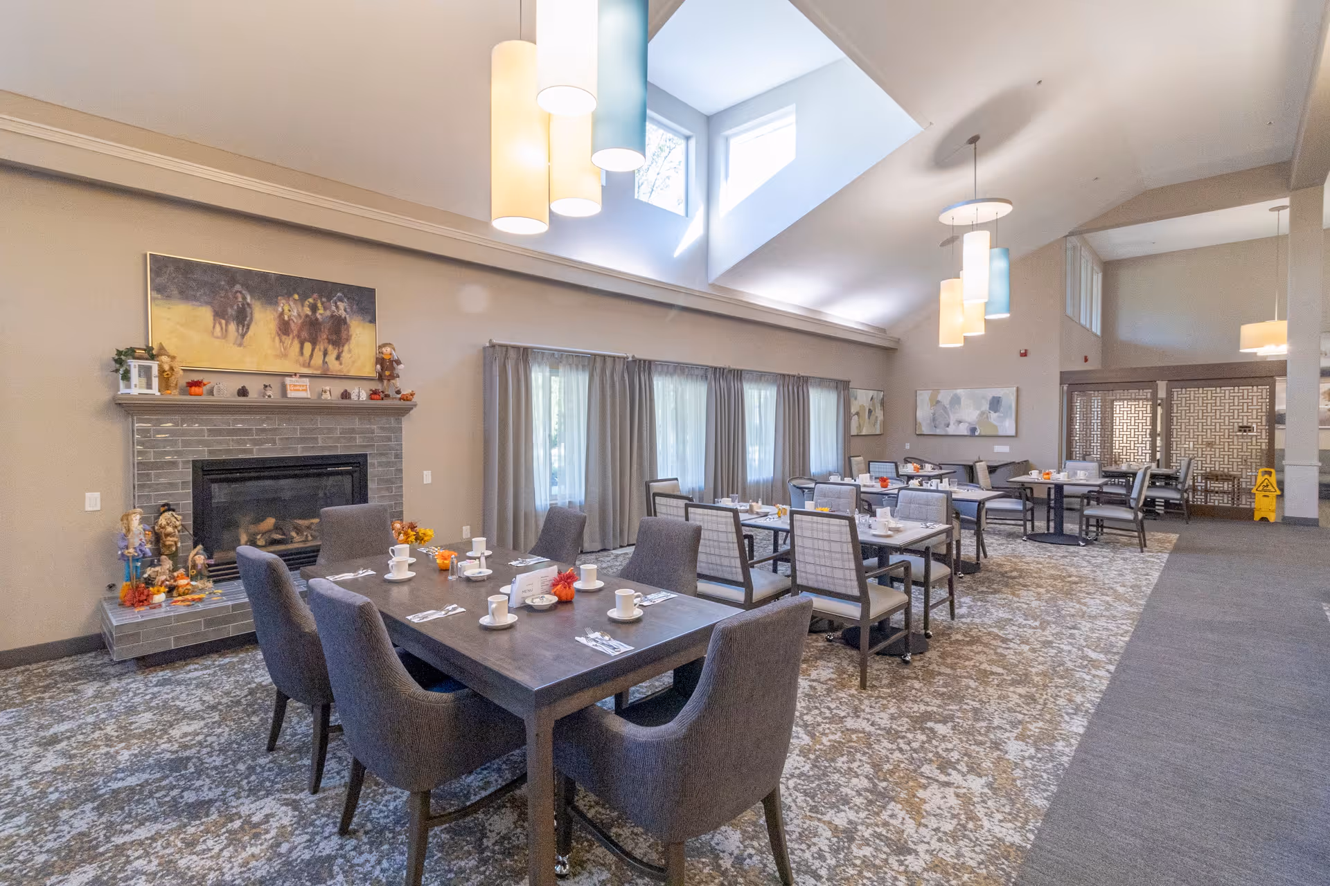 A spacious dining room with several tables set with white cups, plates, and silverware. The room features a fireplace with decorative items on the mantle, large windows with curtains, modern hanging light fixtures, and artwork on the walls. The carpet has a patterned design, and the ceiling is vaulted with skylights allowing natural light to enter.