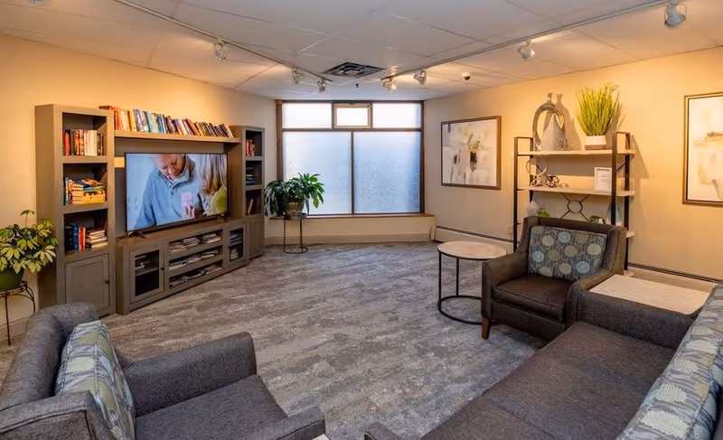 A cozy living room with gray upholstered sofas and armchairs arranged around a small round table. A large entertainment center with a TV and bookshelves is against one wall. The room has a large frosted window, potted plants, abstract artwork on the walls, and warm lighting from ceiling track lights.
