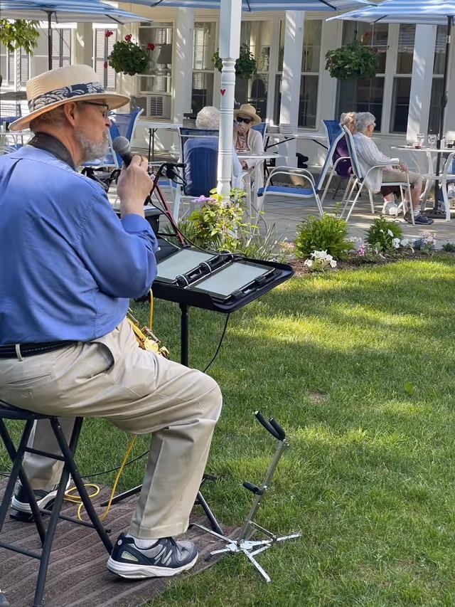 A man wearing a straw hat sings into a microphone outdoors while elderly residents sit on a shaded patio watching him.