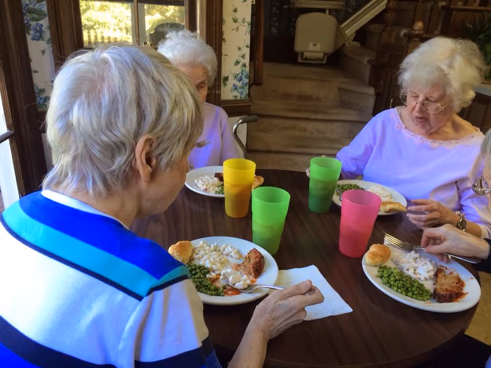 Four elderly women sitting around a round wooden table eating a meal consisting of peas, macaroni and cheese, a bread roll, and a piece of meatloaf. Each has a colorful plastic cup in front of them. The setting appears to be a cozy dining area with floral wallpaper and a staircase in the background.