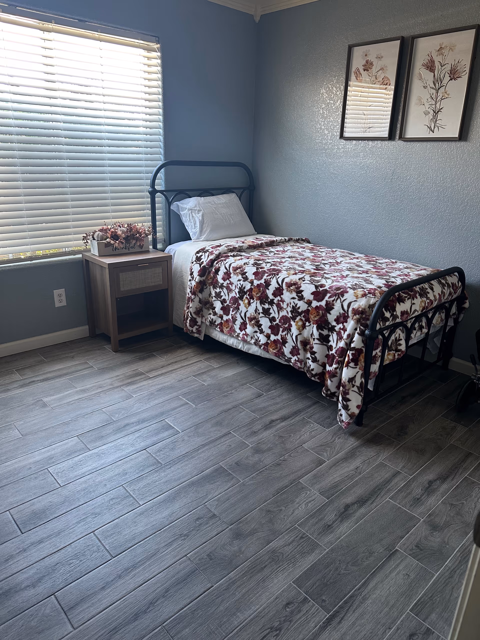 A small bedroom with a single metal-framed bed covered with a floral blanket. Next to the bed is a wooden nightstand with a decorative flower arrangement. The room has gray walls, two framed floral pictures hanging above the bed, and a window with white blinds letting in natural light. The floor is covered with gray wood-like tiles.