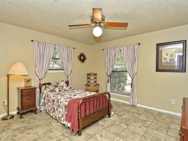 A bedroom with a wooden bed, floral bedding, windows with striped curtains, and a ceiling fan.