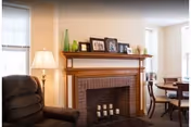 A cozy living area with a brick fireplace and wooden mantel topped with framed photos and vases, an armchair to the left and a dining table to the right.