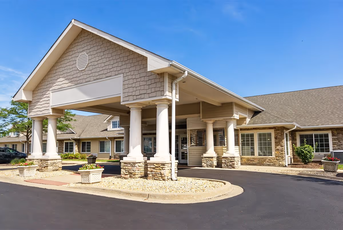 Exterior view of Rock Run Memory Care facility showing the entrance with a covered driveway supported by white columns with stone bases, beige siding, and multiple windows under a clear blue sky.