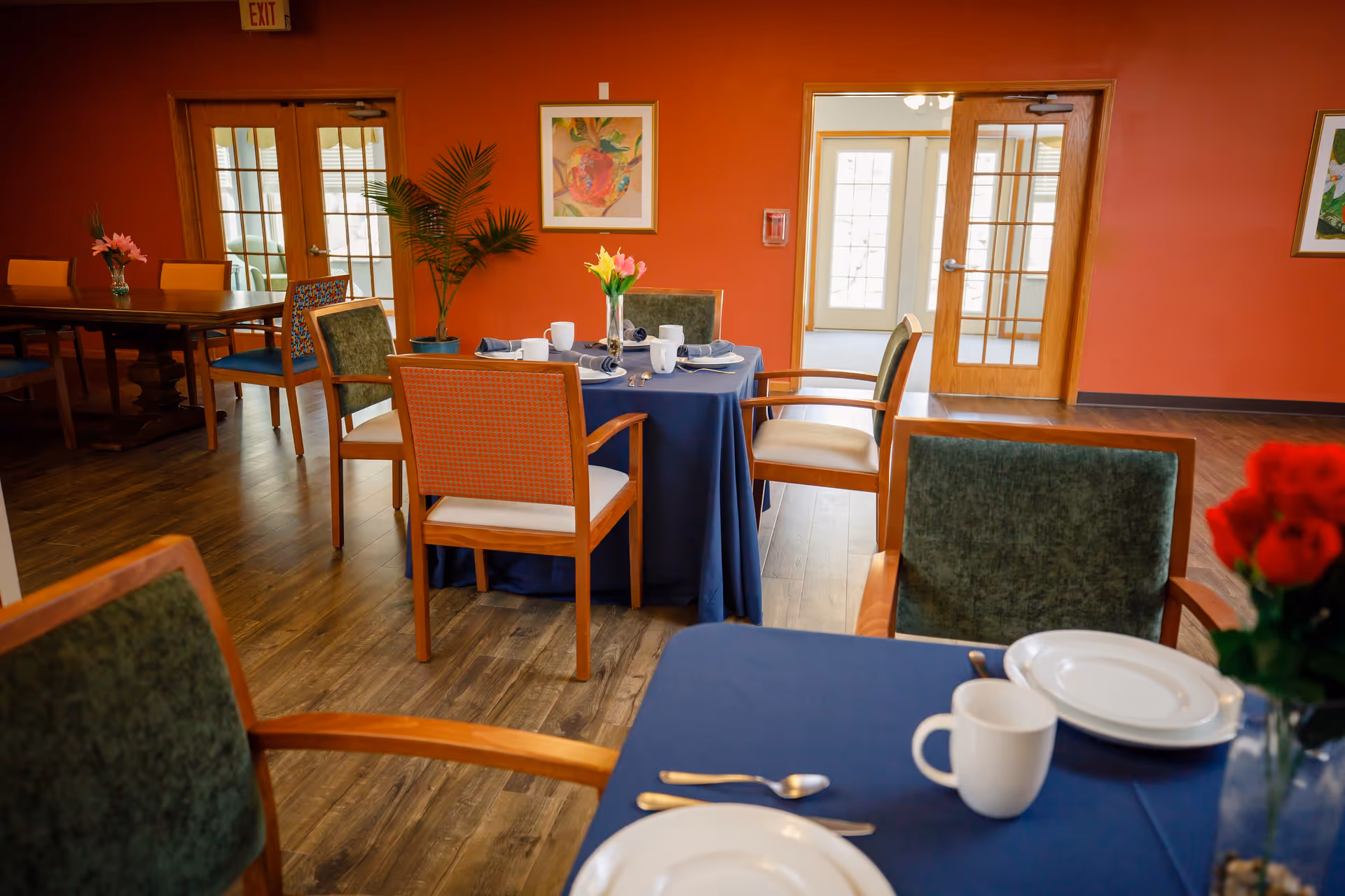 A dining room with wooden chairs and tables covered with blue tablecloths. The tables are set with white plates, cups, silverware, and napkins. There are colorful flowers in vases on the tables. The room has orange walls, wooden flooring, framed artwork, and glass-paneled wooden doors leading to another area.