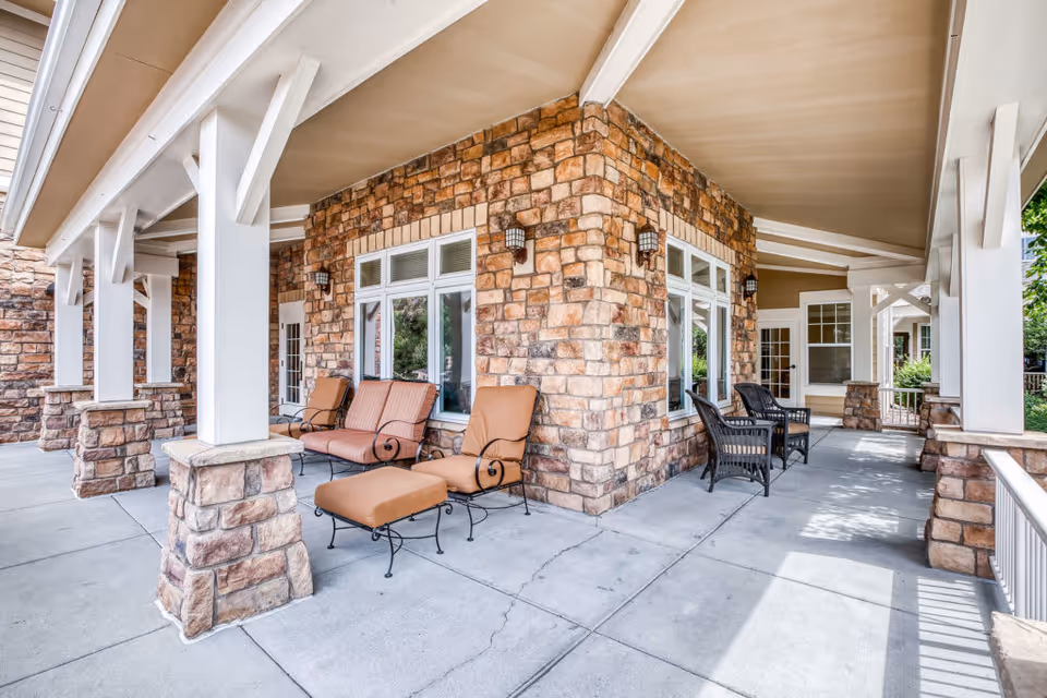 Covered outdoor patio area with stone pillars and walls, featuring cushioned chairs and a loveseat, along with black wicker chairs. The space is shaded by a beige ceiling and has large windows and doors leading inside.