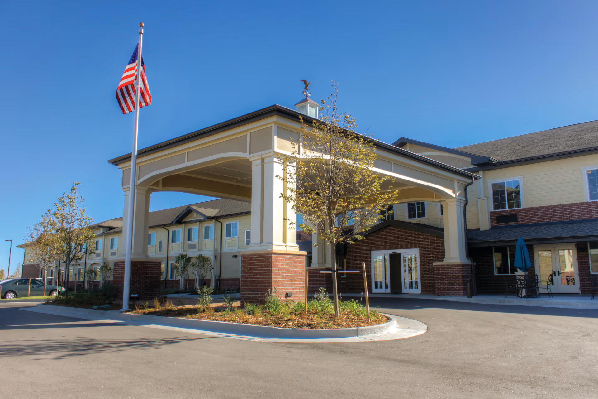 Exterior view of Peakview Assisted Living and Memory Care facility showing the main entrance with a covered drop-off area, an American flag on a flagpole, small trees, and a clear blue sky.