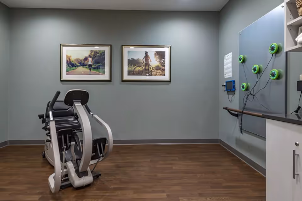 Small exercise/therapy room with a recumbent exercise machine, two framed photos on the wall, and a panel with suction devices on the right.