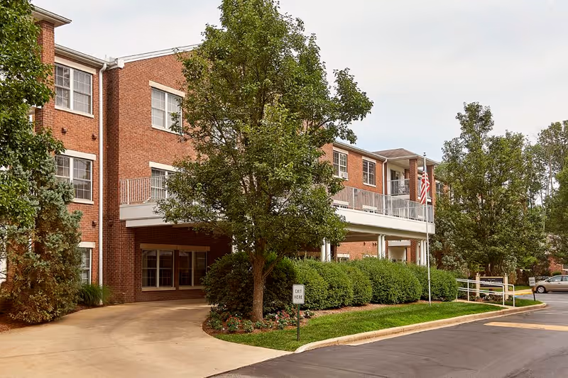 Exterior view of a three-story brick building with multiple windows, surrounded by trees and bushes. There is a driveway leading to a covered entrance area, an American flag on a flagpole, and a sign that reads 'EXIT HERE'.