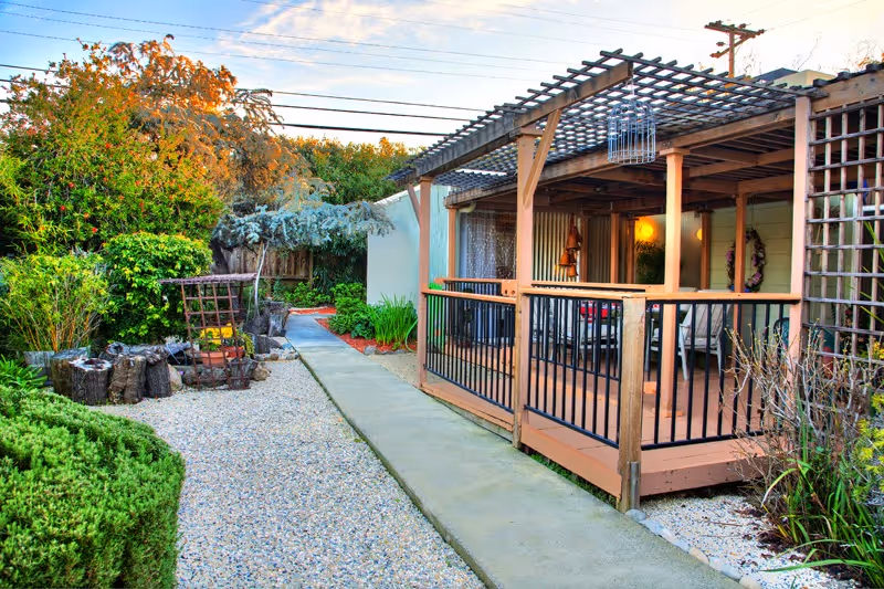 A landscaped outdoor courtyard featuring a pergola-covered porch with seating, a concrete walkway and gravel beds surrounded by shrubs.