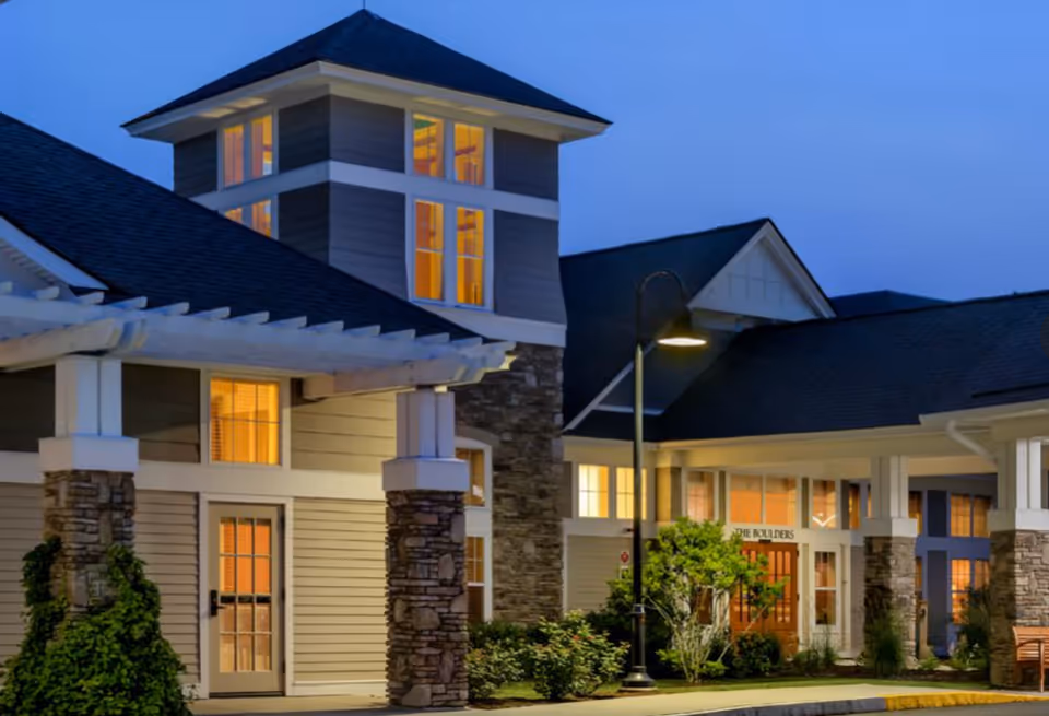 Exterior view of a senior living facility building named The Boulders at dusk, featuring stone pillars, lit windows, and a street lamp illuminating the entrance area.