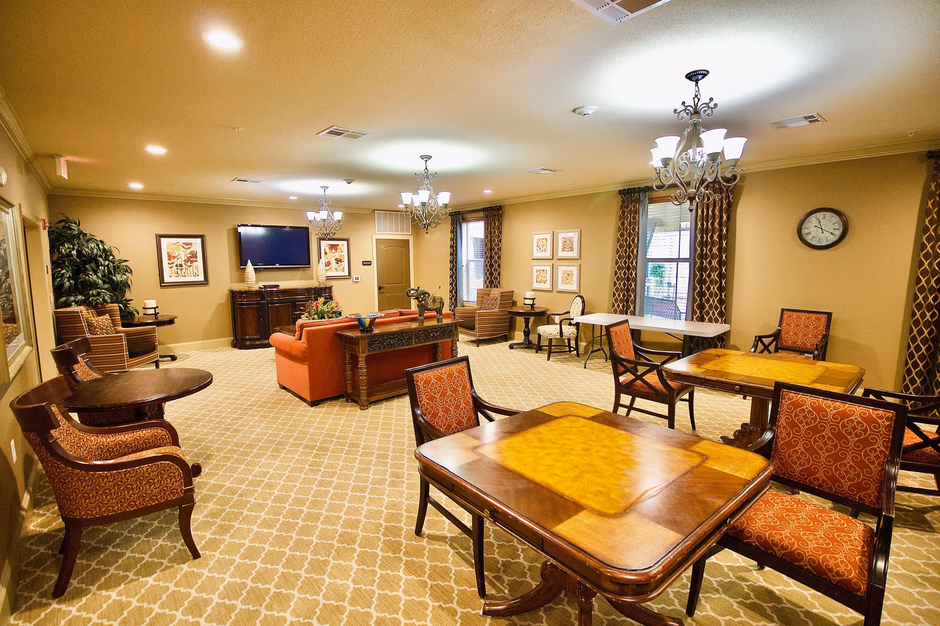 Bright, well-furnished senior living common room with multiple tables and chairs, sofas, a wall-mounted TV, chandeliers and patterned curtains.