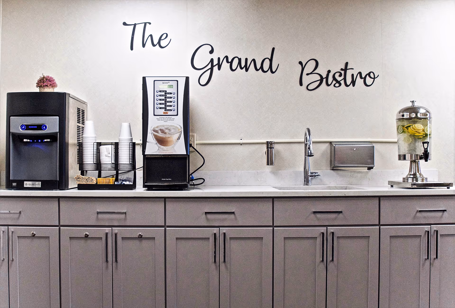 Countertop bistro station with coffee machines, cup dispensers, a sink and a lemon water dispenser beneath a wall sign that reads "The Grand Bistro".